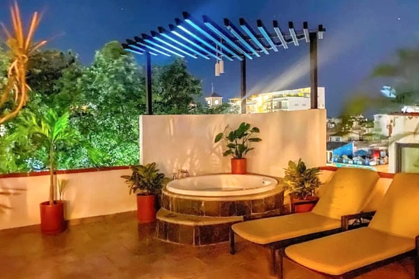 Rooftop terrace at night with illuminated pergola, circular hot tub, potted plants, lounge chairs, and city views