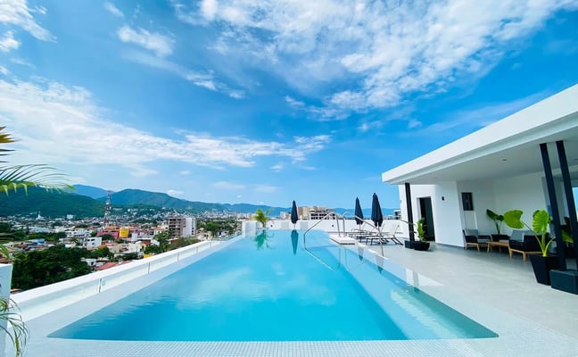 Rooftop infinity pool with city and mountain views, white modern building, lounge chairs with umbrellas