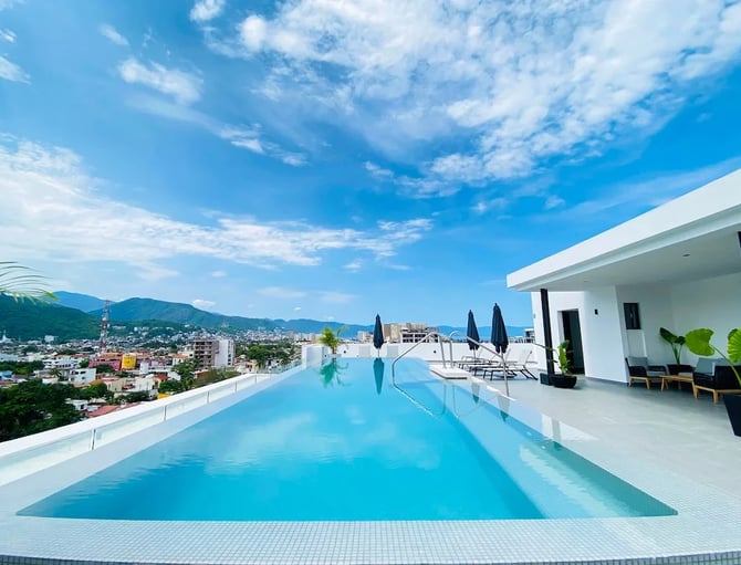 Rooftop infinity pool with city and mountain views, white modern building, lounge chairs with umbrellas